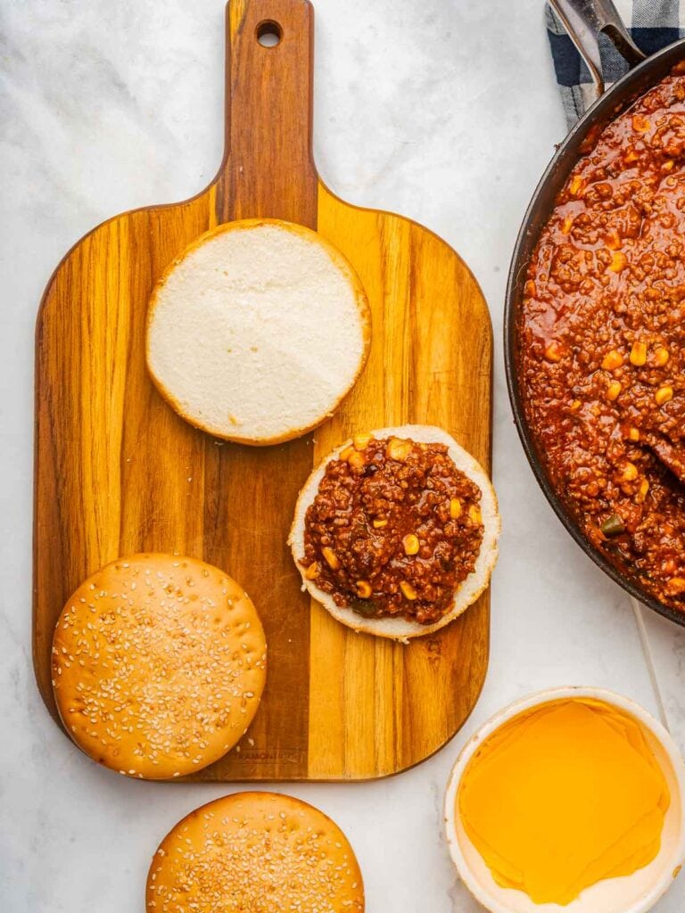 Toasted buns on a cutting board being assembled with the taco meat mixture and cheese slices nearby.