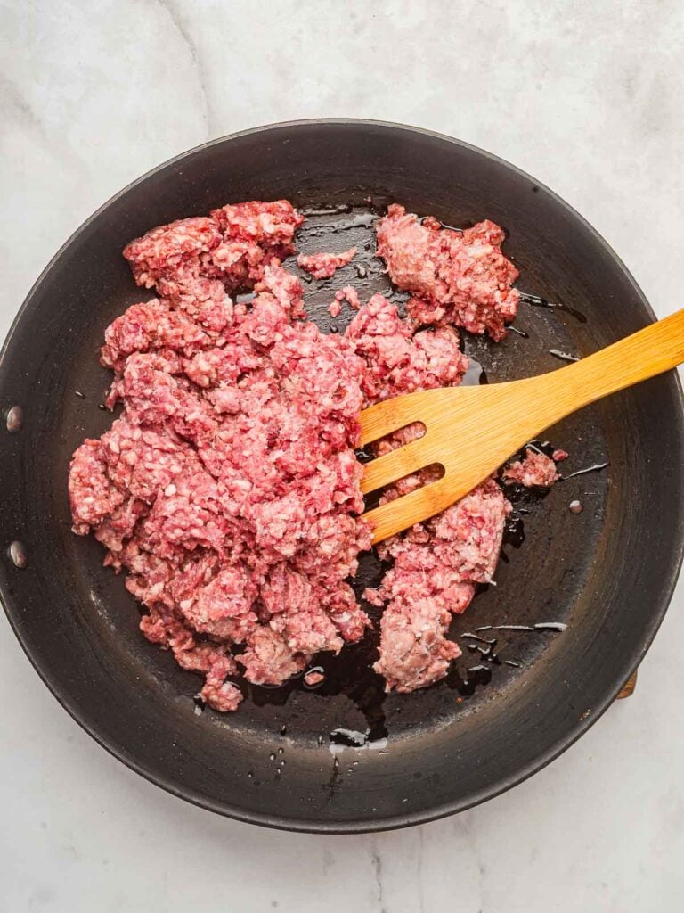 Raw ground beef in a skillet starting to brown with oil and a wooden spatula resting in the pan.