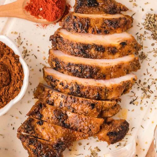 Sliced chipotle chicken on a white board with spices, garlic, and herbs scattered around.
