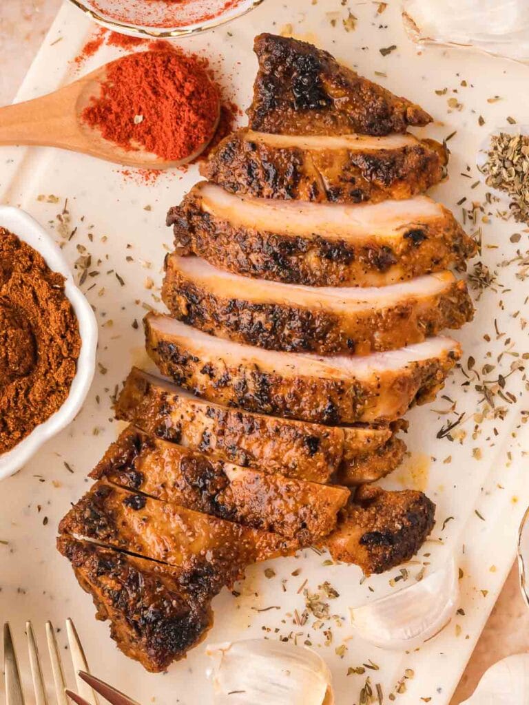 Sliced chipotle chicken on a white board with spices, garlic, and herbs scattered around.