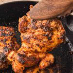 Juicy chipotle chicken thighs seared in a cast iron skillet with a wooden spoon lifting one out.