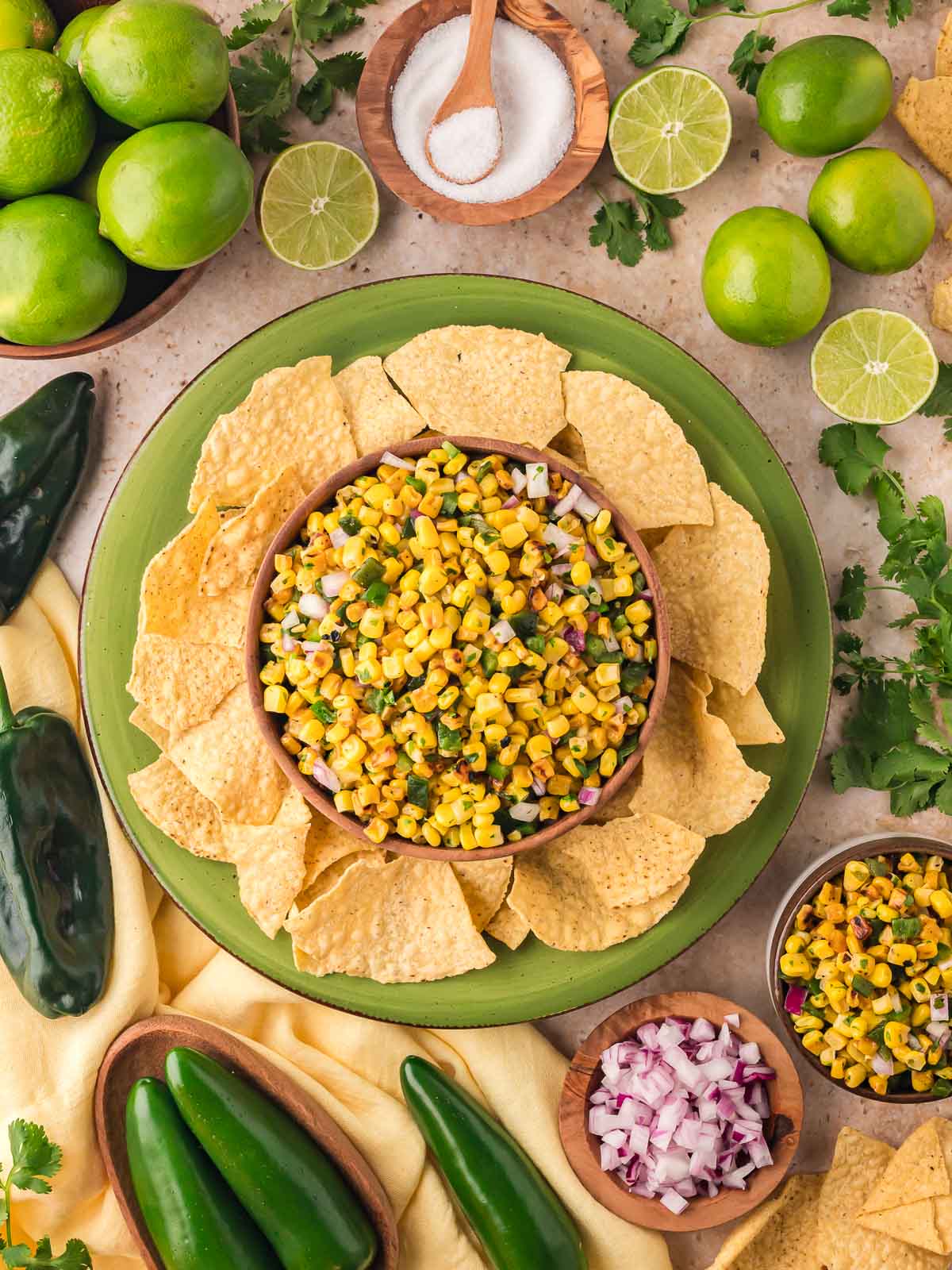 Top-down view of a bowl of Chipotle-style corn salsa surrounded by tortilla chips, fresh limes, poblano peppers, and cilantro.