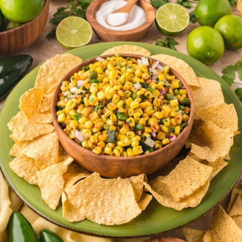 Front view of a bowl of Chipotle-style corn salsa surrounded by tortilla chips, fresh limes, poblano peppers, and cilantro.