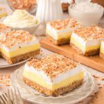 Coconut cream bar on a small plate with more bars in the background, surrounded by coconut shells, graham crackers, and toasted coconut.