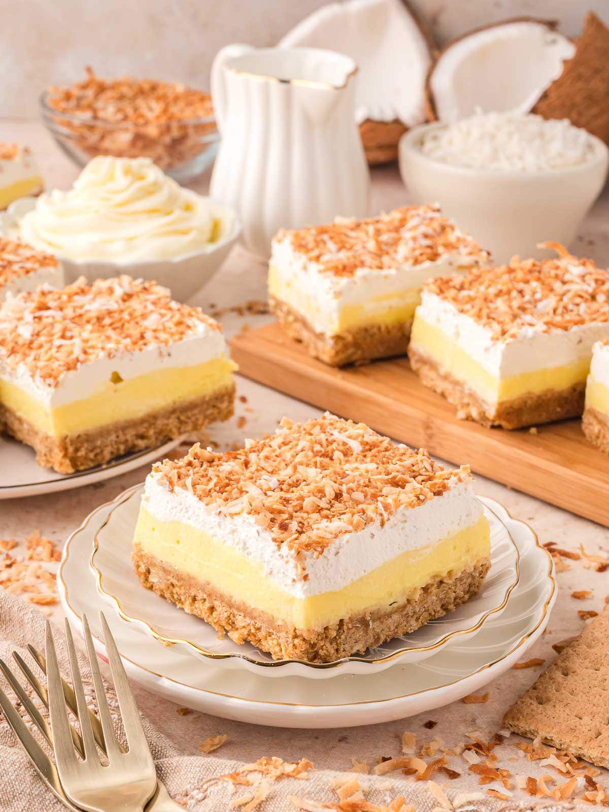 Coconut cream bar on a small plate with more bars in the background, surrounded by coconut shells, graham crackers, and toasted coconut.