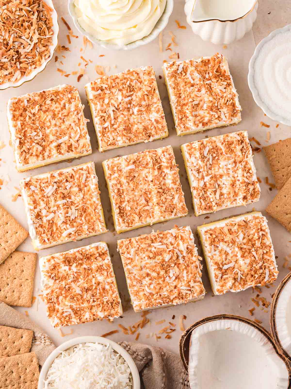 Nine coconut cream bars topped with toasted coconut, surrounded by graham crackers, shredded coconut, whipped topping, and coconut halves.