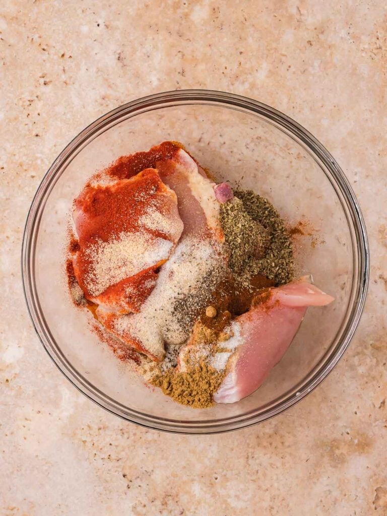 Raw chicken thighs in a bowl with chipotle seasoning and spices before mixing