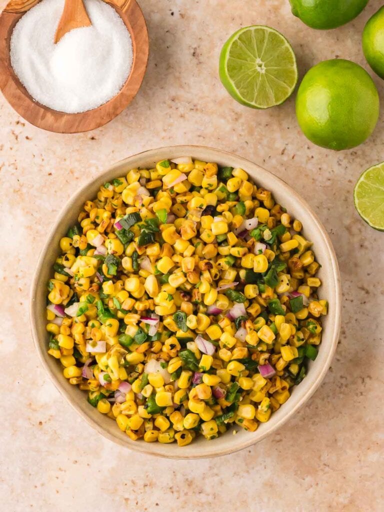 Overhead shot of a finished bowl of roasted corn salsa with lime halves, salt, and fresh cilantro styled around it, ready to serve.