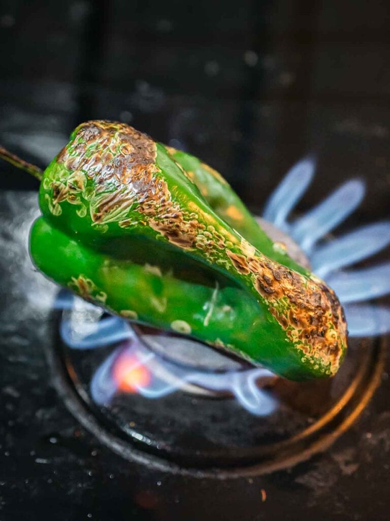 Poblano pepper being charred over an open flame on a stovetop, showing the roasting step for Chipotle corn salsa.