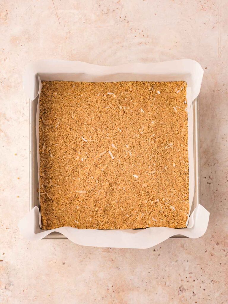 Graham cracker and coconut crust pressed into a parchment-lined 9x9 pan, ready to chill.