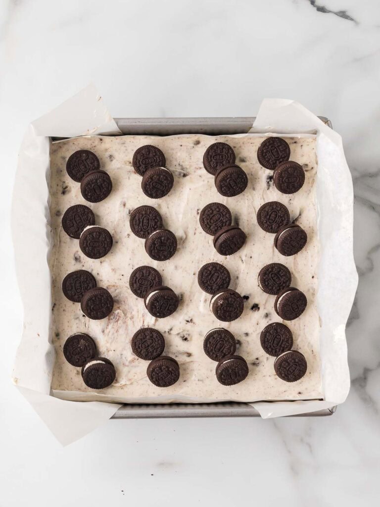 Mini Oreos arranged on top of the cookies and cream cheesecake layer in a parchment-lined brownie pan before chilling.