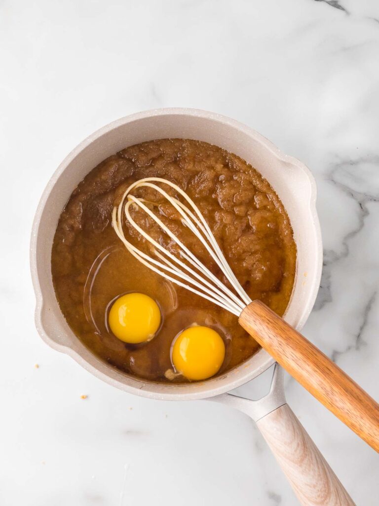 Two eggs just added to a saucepan of melted butter and sugar with a whisk resting inside.