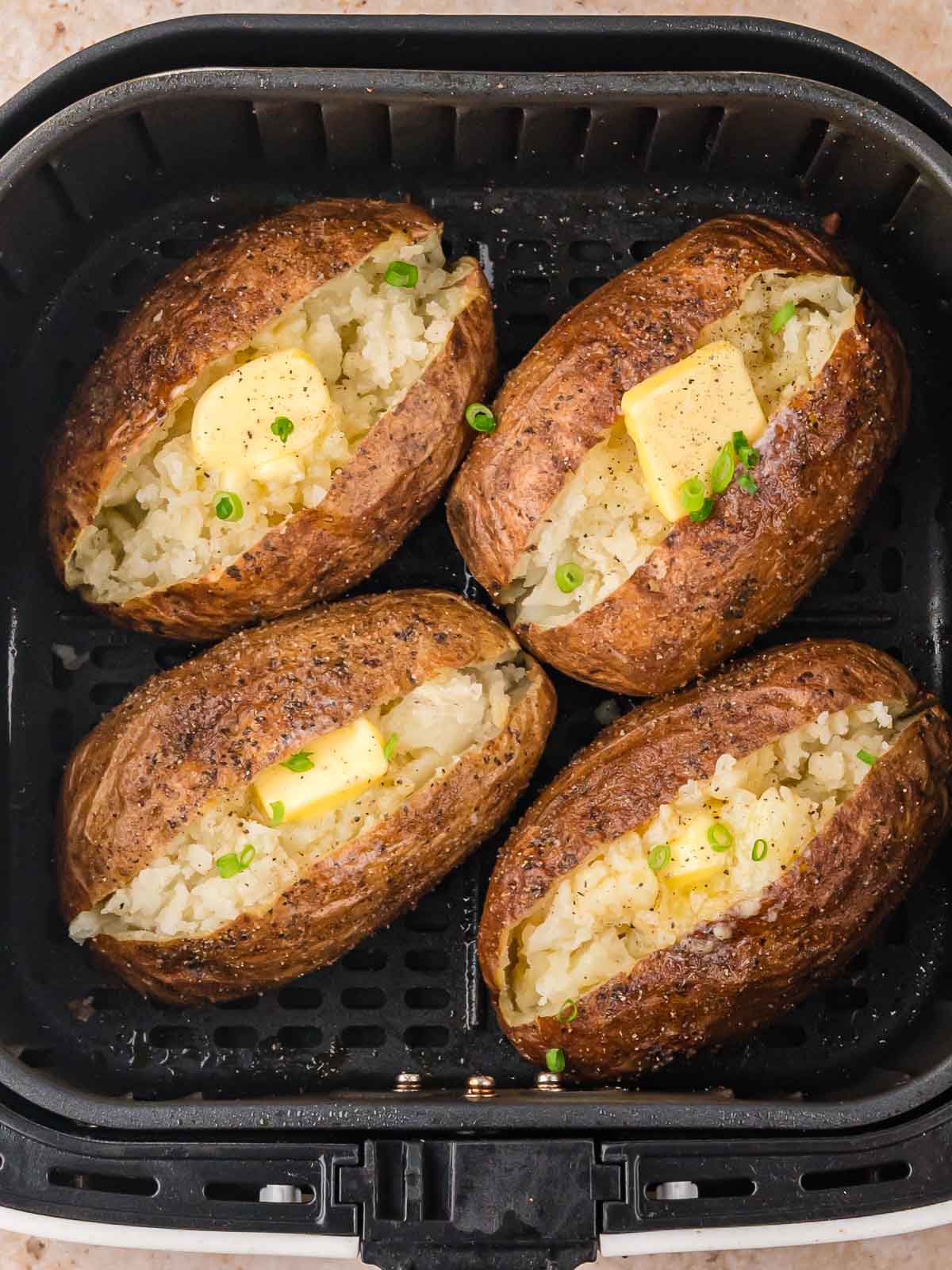 Four baked potatoes in an air fryer basket, split open and topped with butter and chopped green onions.