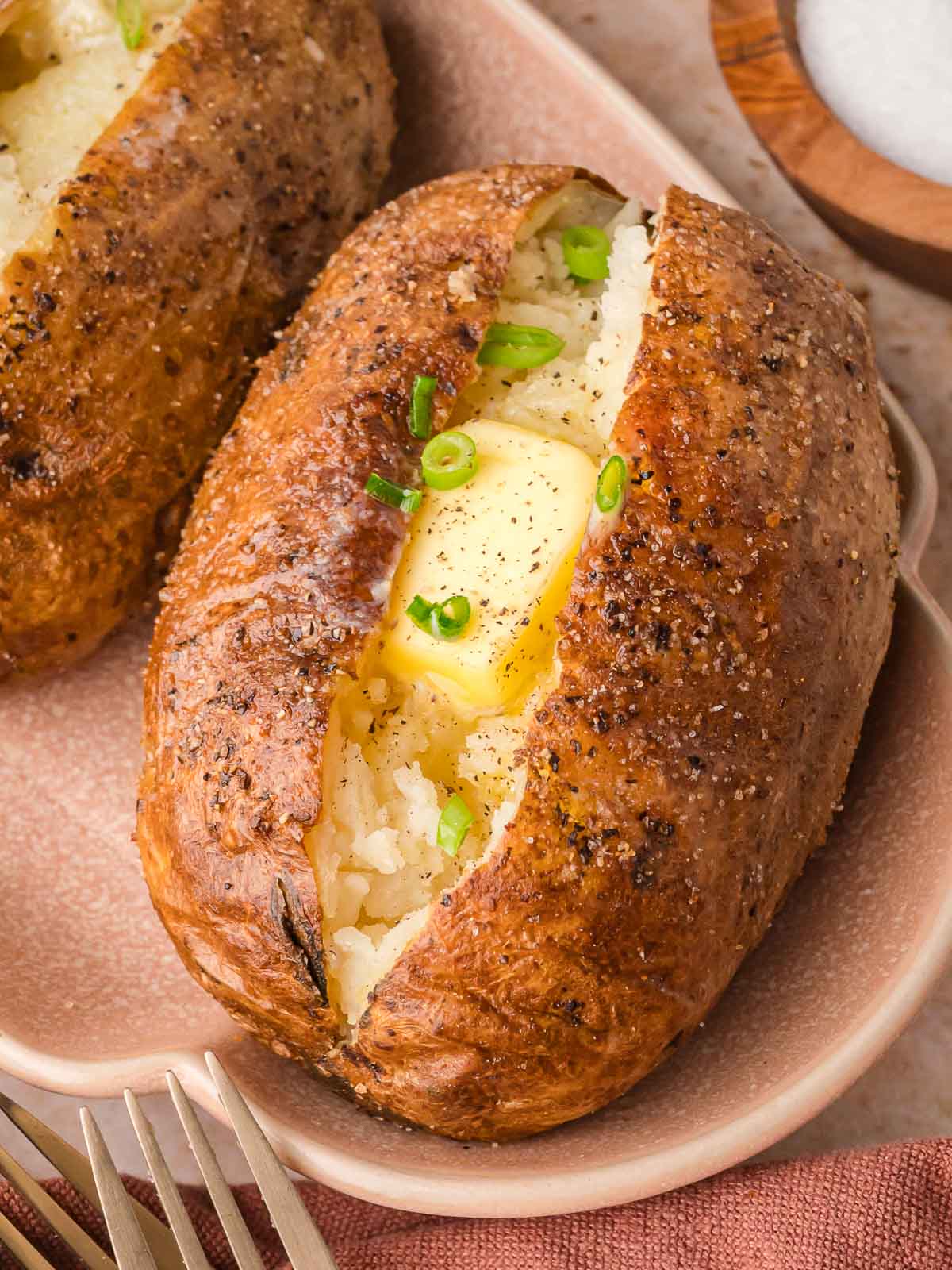 Close-up of a baked potato on a pink plate, with melted butter and green onions on top of a fluffy center.