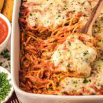 Baked spaghetti casserole topped with melted cheese and parsley, served in a white baking dish with a wooden spoon.