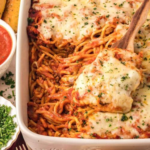 Baked spaghetti casserole topped with melted cheese and parsley, served in a white baking dish with a wooden spoon.