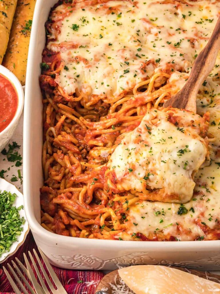 Baked spaghetti casserole topped with melted cheese and parsley, served in a white baking dish with a wooden spoon.