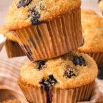Two tall blueberry muffins stacked on a gingham cloth with coarse sugar and berries scattered around.