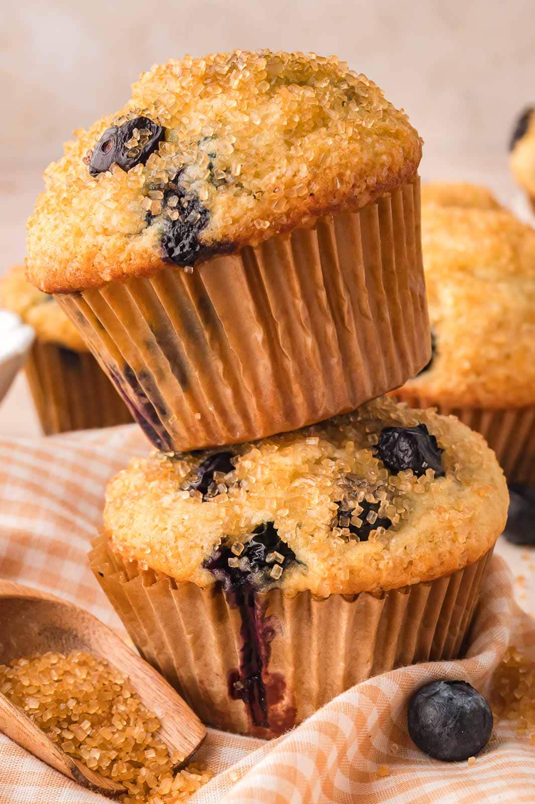 Two tall blueberry muffins stacked on a gingham cloth with coarse sugar and berries scattered around.