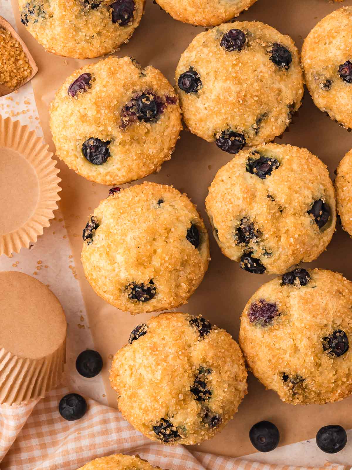 A batch of golden blueberry muffins with crunchy sugar tops on parchment, surrounded by fresh blueberries and muffin liners.