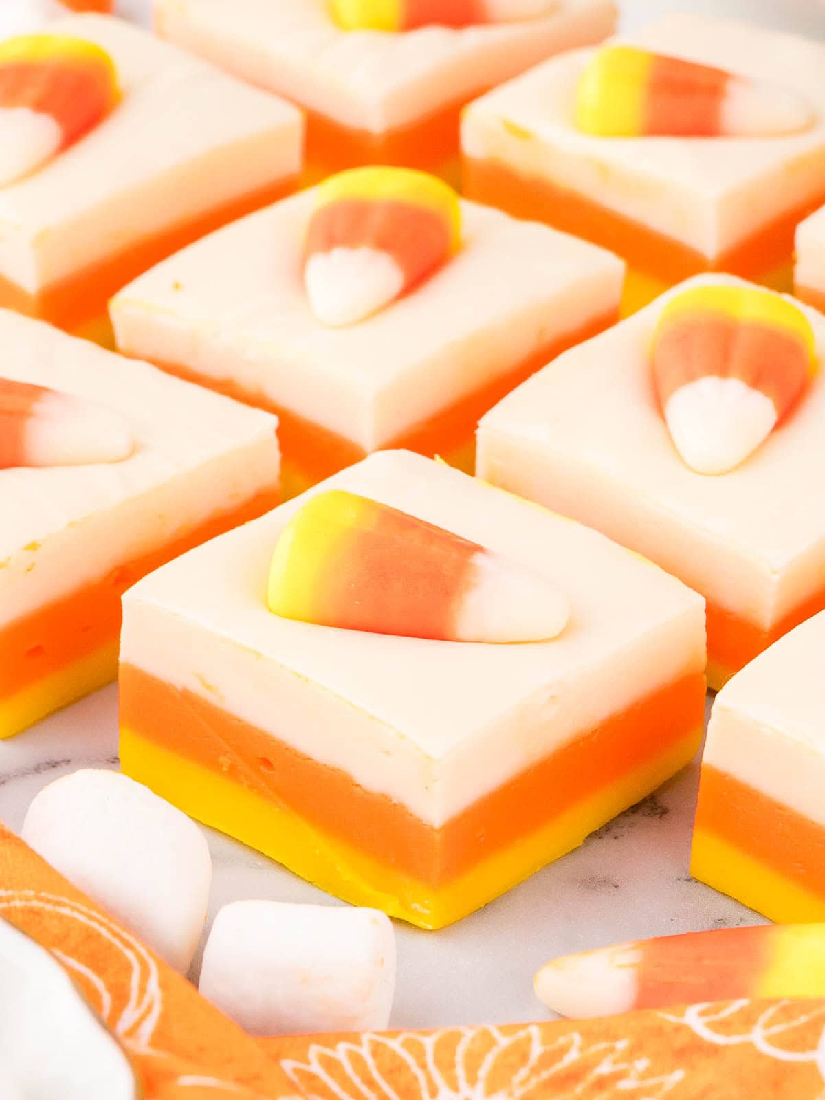 Close-up of neat fudge squares topped with candy corn, arranged in rows on a marble surface with orange napkin accents.