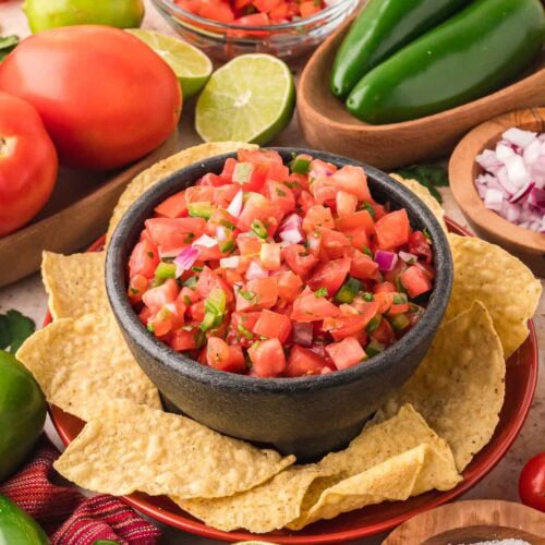 Fresh tomato salsa served in a black bowl with tortilla chips, surrounded by limes, jalapeños, and tomatoes.