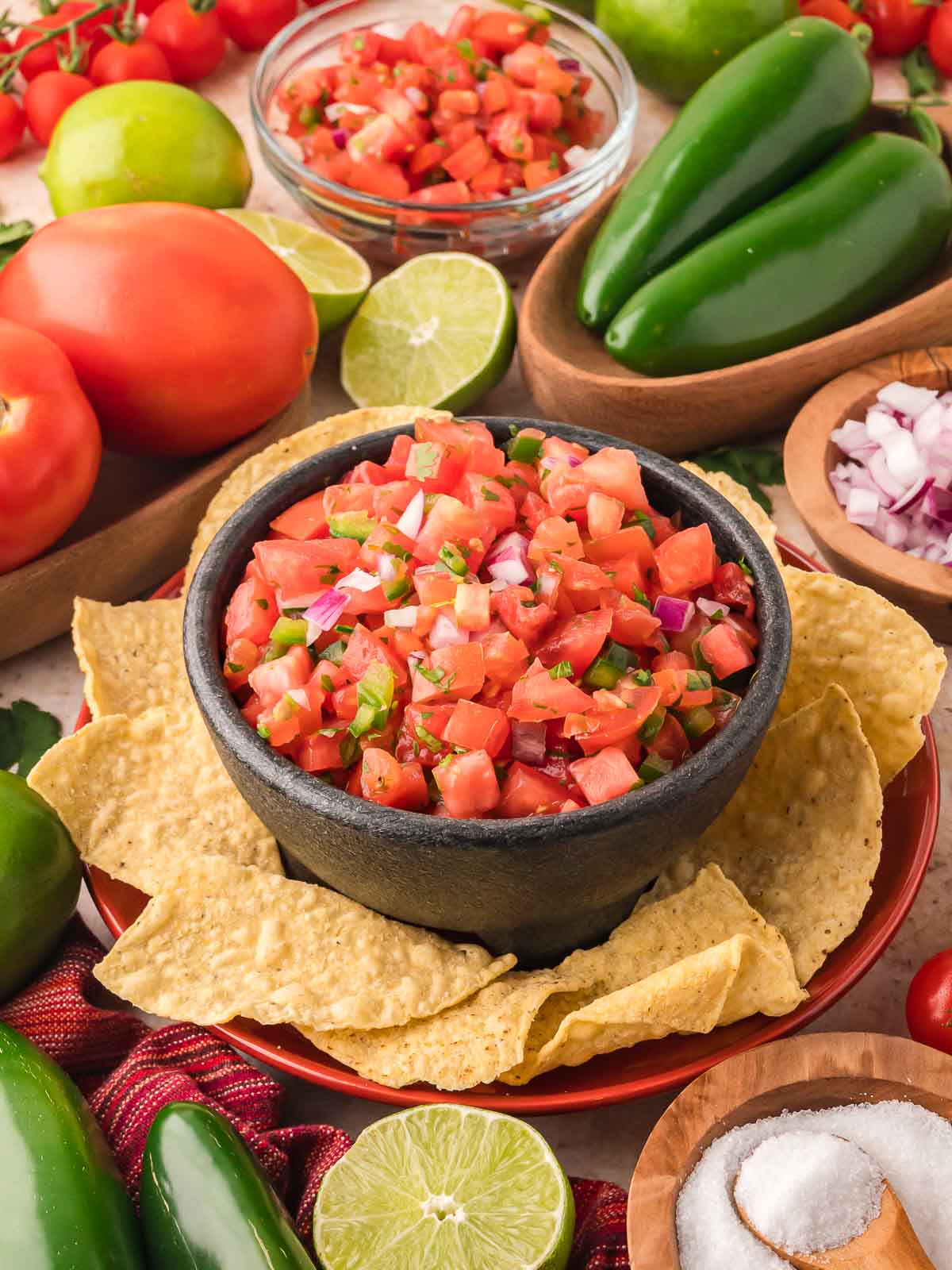 Fresh tomato salsa served in a black bowl with tortilla chips, surrounded by limes, jalapeños, and tomatoes.