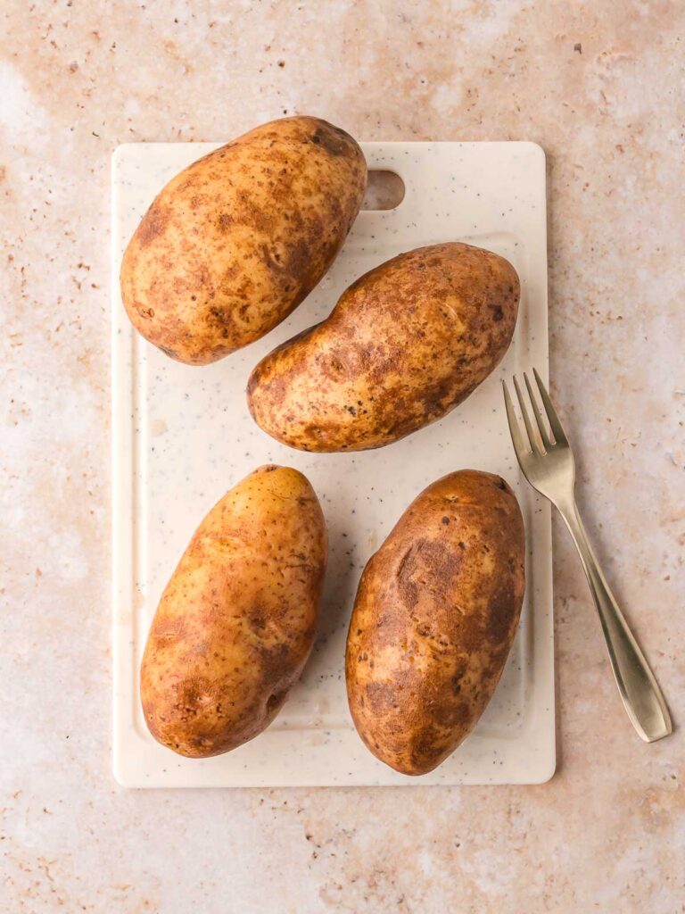 Four raw russet potatoes on a white cutting board with a fork, ready to be prepped.