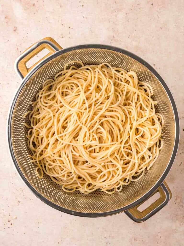 Cooked spaghetti noodles drained in a metal colander over a light countertop.