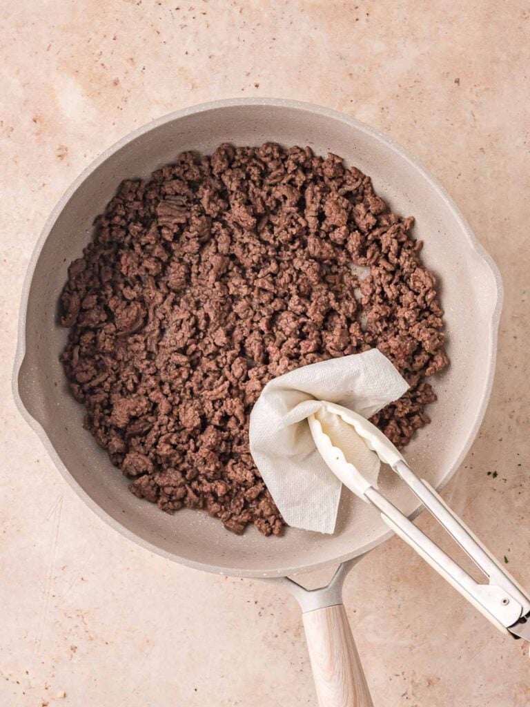 Ground beef fully browned in a skillet with grease being blotted out using a paper towel and tongs.