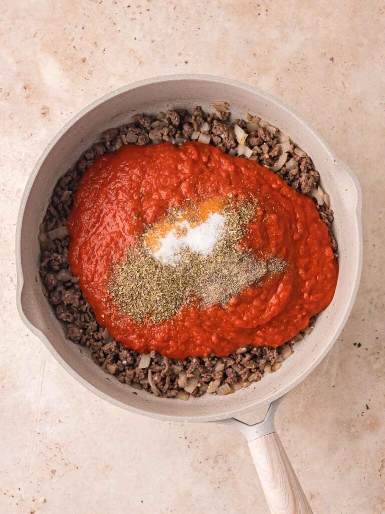 Marinara sauce, herbs, salt, and pepper added on top of the cooked beef and onions in the pan.