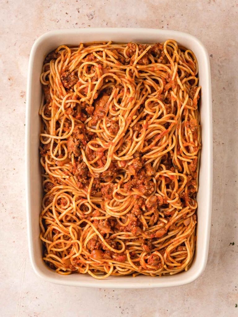 Cooked spaghetti tossed with meat sauce and placed into a rectangular baking dish before adding cheese.