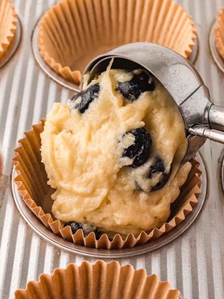 Muffin batter with blueberries being scooped into a lined muffin tin.