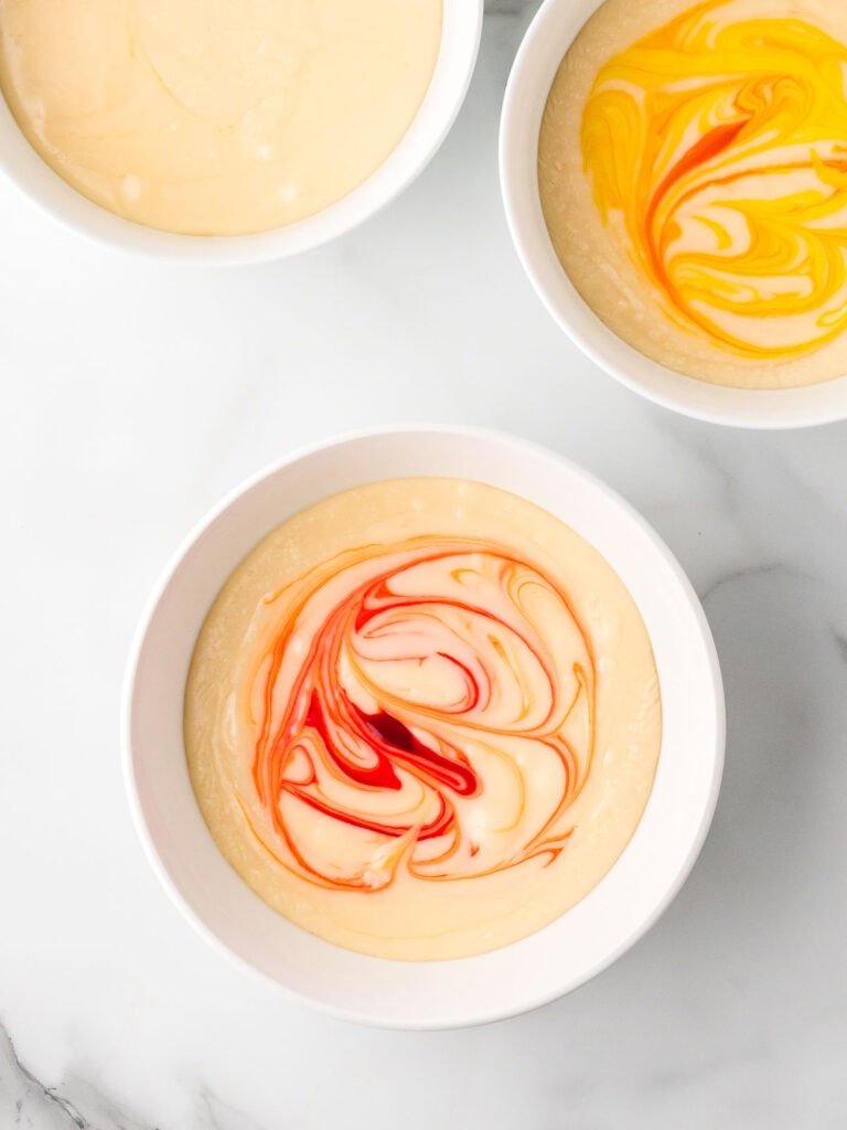 Three bowls of melted fudge mixture, one plain and two swirled with yellow and orange food coloring, ready to be stirred.