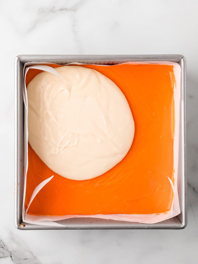 White fudge layer poured over the orange layer, almost ready to be smoothed out as the final layer.
