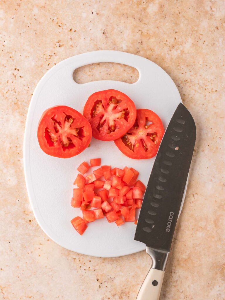 Sliced and diced Roma tomatoes on a white cutting board with a kitchen knife on a beige countertop.