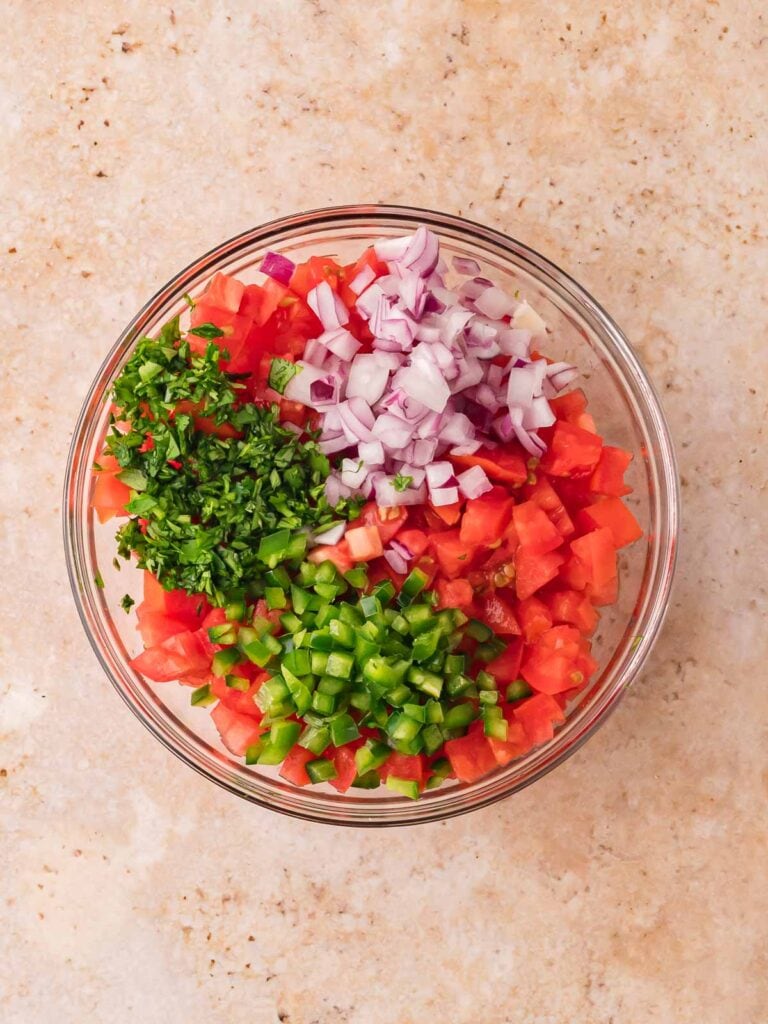 Glass bowl filled with chopped Roma tomatoes, red onion, jalapeño, and cilantro before mixing.
