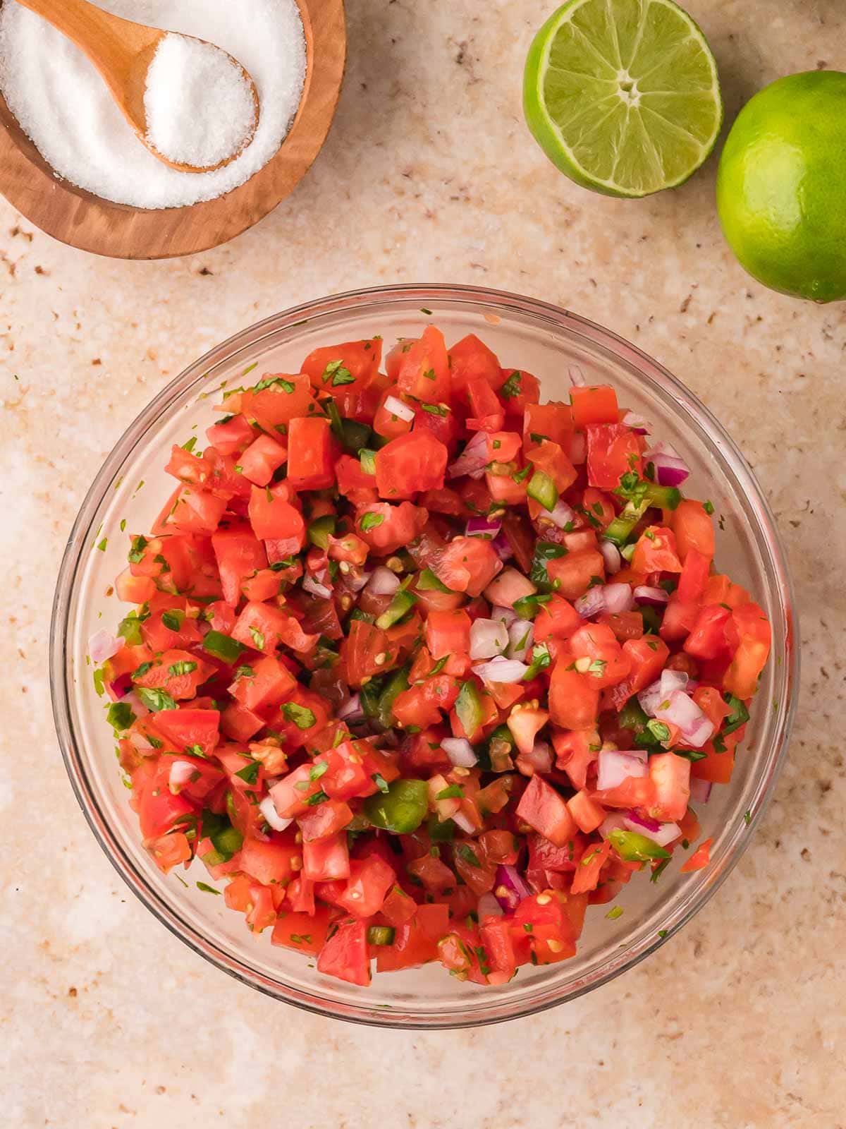 Fresh tomato salsa in a bowl surrounded by lime halves and a wooden bowl of kosher salt.