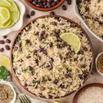 Overhead shot of a serving bowl of black beans and rice with lime slices, surrounded by small bowls of ingredients.