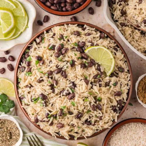 Overhead shot of a serving bowl of black beans and rice with lime slices, surrounded by small bowls of ingredients.