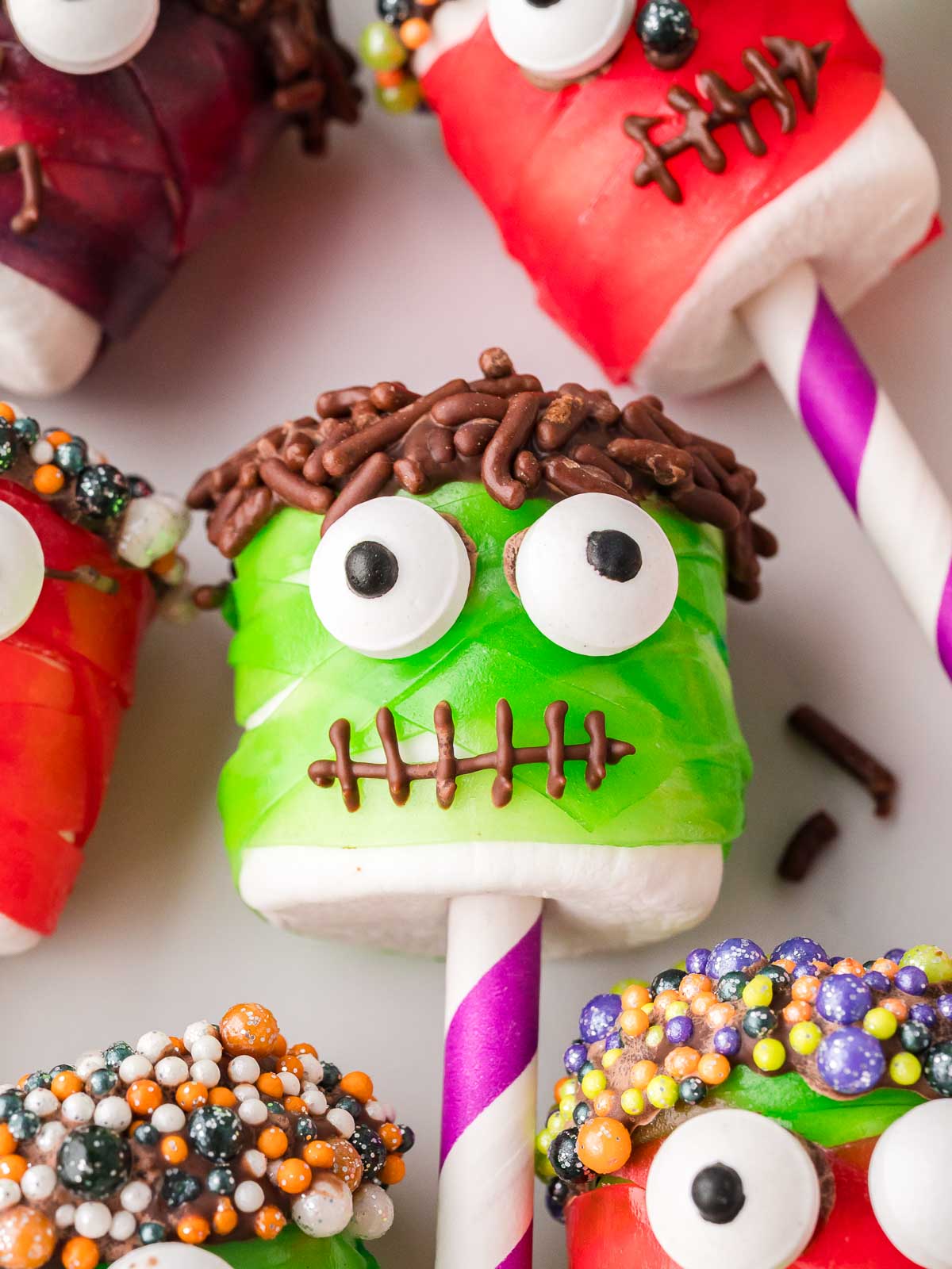 Green marshmallow monster pop on a purple straw decorated with candy eyes, chocolate stitches, and chocolate sprinkle hair.