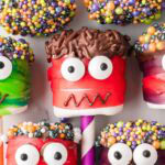Colorful marshmallow monster pops with red and green fruit roll-up bandages, candy eyeballs, and sprinkle-covered chocolate hair.