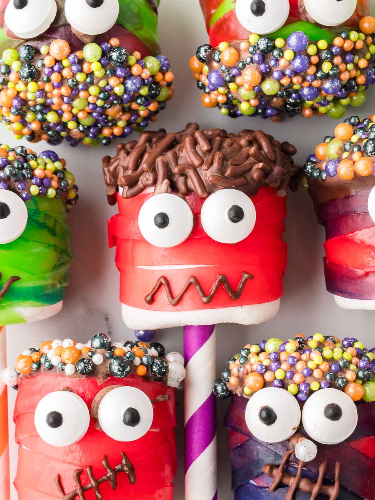 Colorful marshmallow monster pops with red and green fruit roll-up bandages, candy eyeballs, and sprinkle-covered chocolate hair.