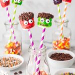 Halloween marshmallow monster pops displayed in glass jars filled with candy corn, surrounded by bowls of sprinkles and candy eyes.