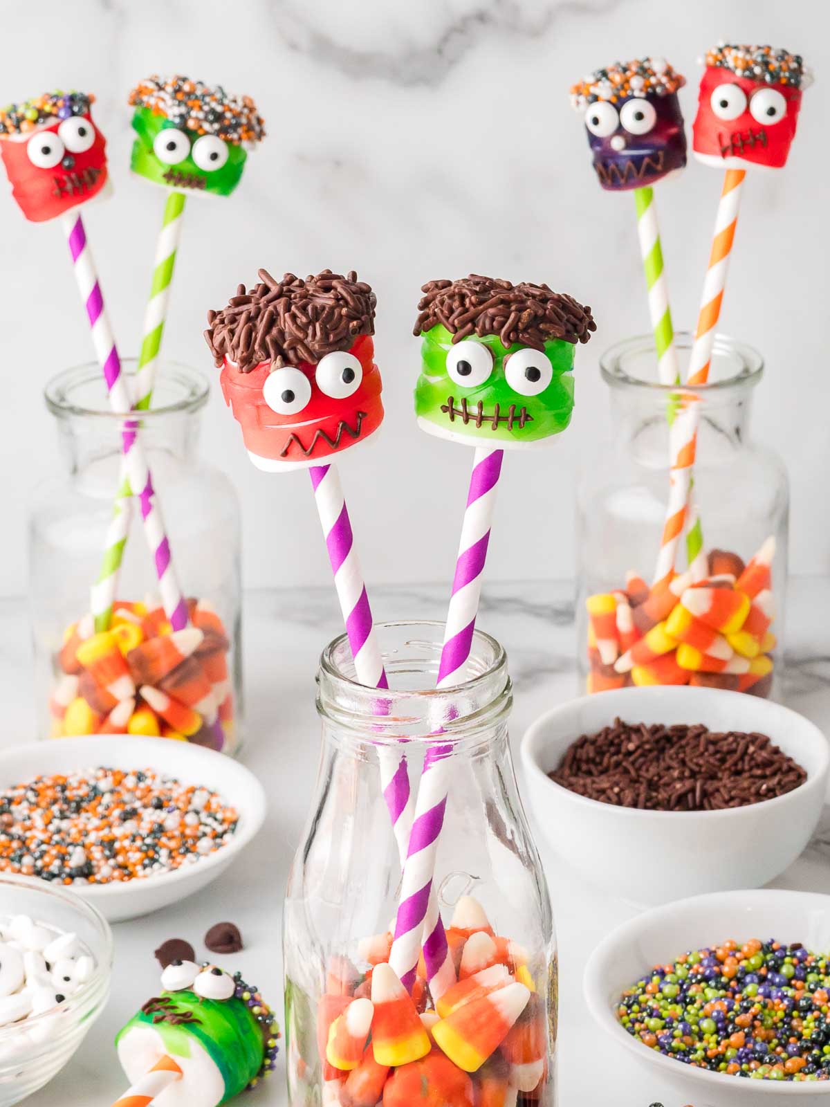 Halloween marshmallow monster pops displayed in glass jars filled with candy corn, surrounded by bowls of sprinkles and candy eyes.