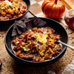 Bowl of pumpkin chili topped with sour cream, shredded cheddar, green onions, and crispy bacon, served with blue corn tortilla chips.