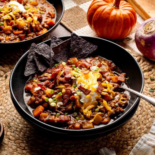 Bowl of pumpkin chili topped with sour cream, shredded cheddar, green onions, and crispy bacon, served with blue corn tortilla chips.