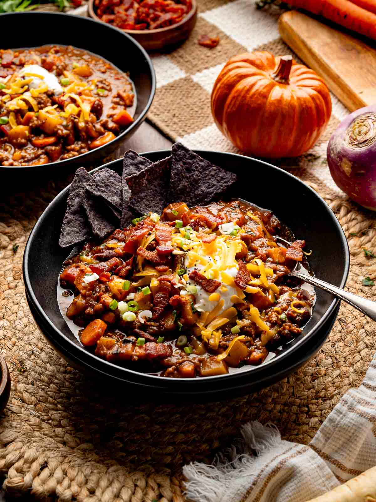 Bowl of pumpkin chili topped with sour cream, shredded cheddar, green onions, and crispy bacon, served with blue corn tortilla chips.