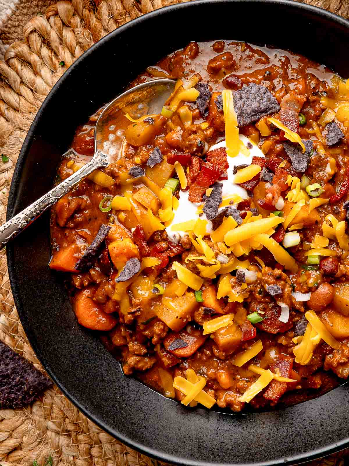 Close-up of loaded pumpkin chili in a black bowl, topped with cheese, bacon, sour cream, and crushed tortilla chips.