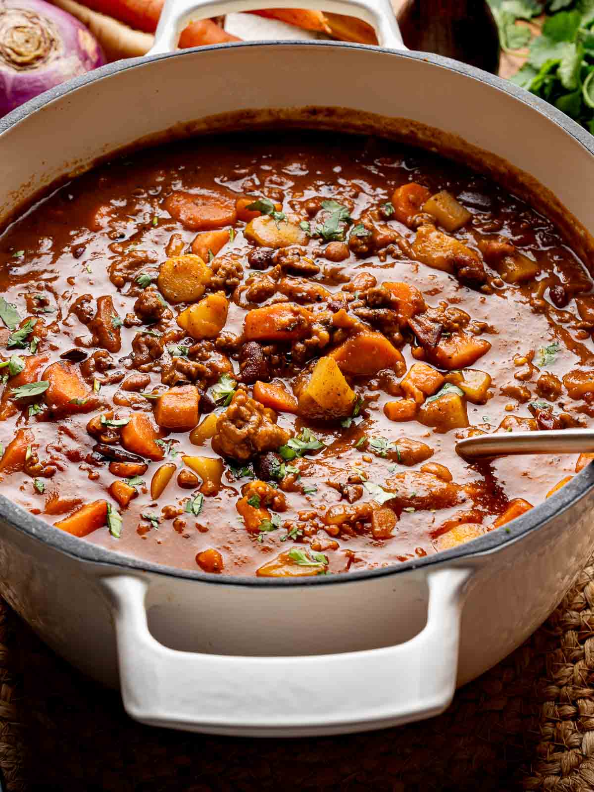 : Overhead shot of a large white pot filled with hearty pumpkin chili made with ground beef, bacon, and chopped root vegetables.